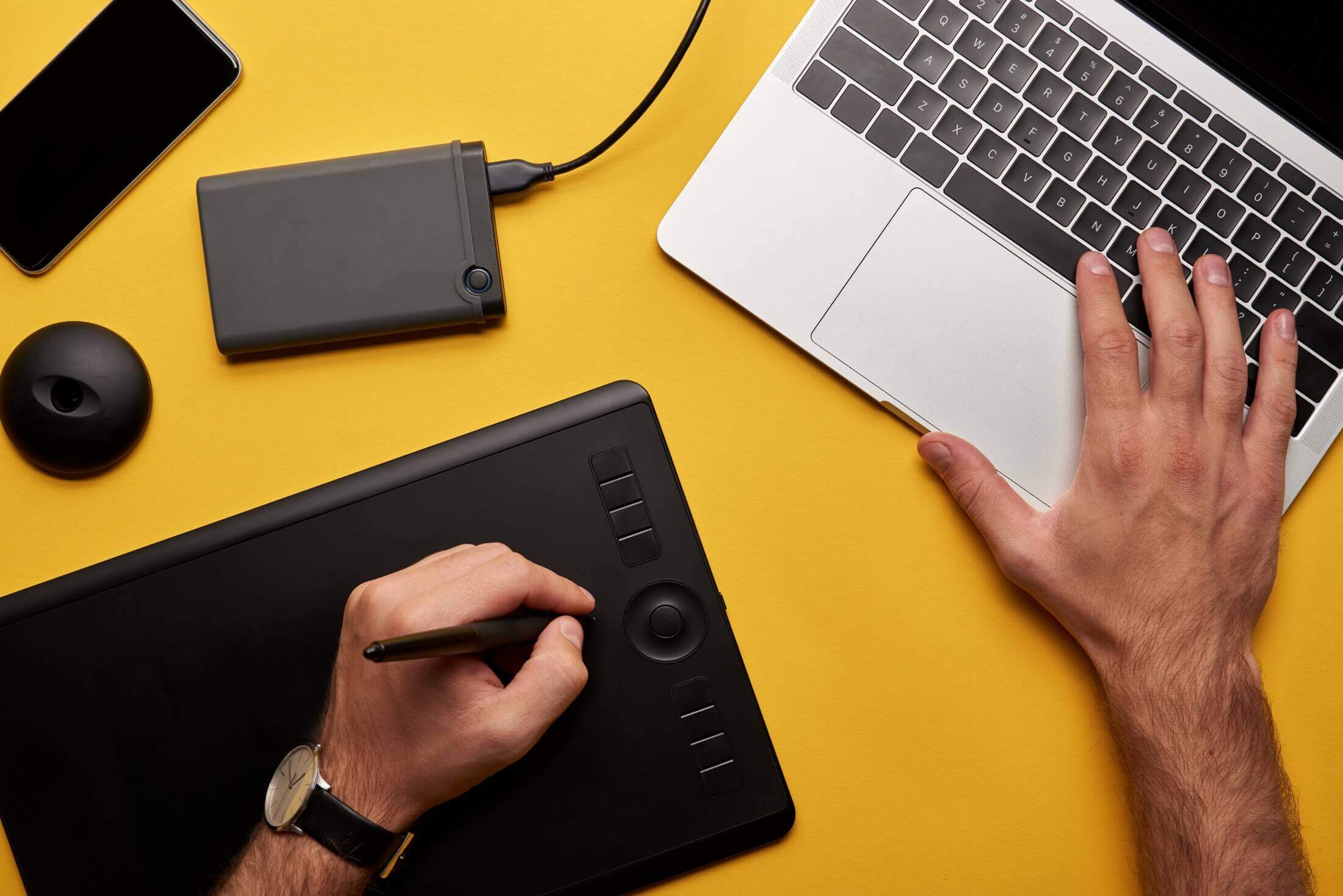 A graphic designer using a stylus pen on a drawing tablet alongside a laptop on a yellow background, with a smartphone and external hard drive also visible.