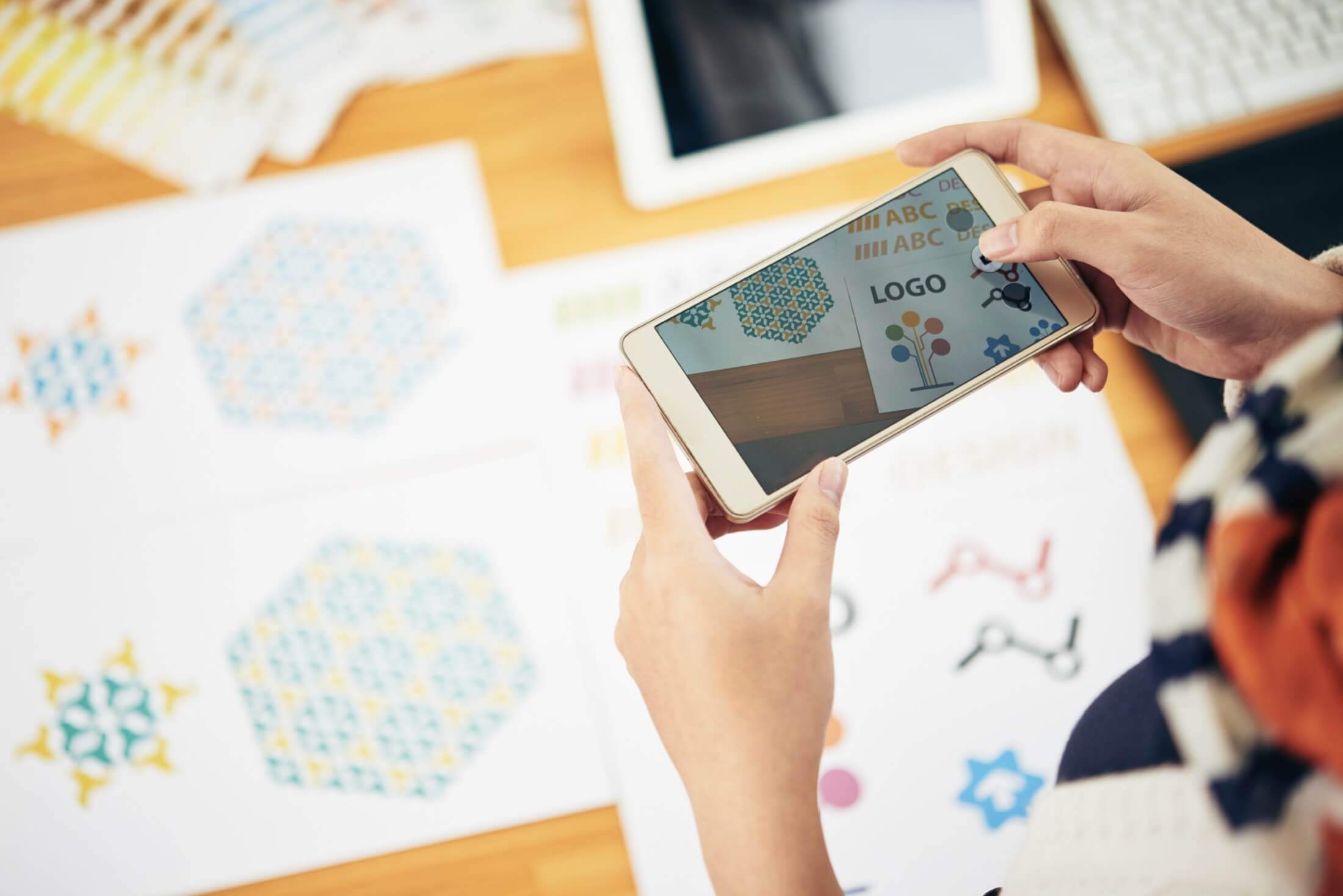A person holding a smartphone displaying various logo designs above a table with scattered printouts of graphic symbols and logo options.