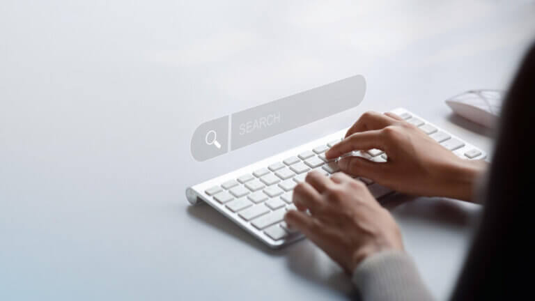 Close-up of a person's hands typing on a white computer keyboard with a transparent 'search' bar graphic overlay, symbolizing online search and digital research.