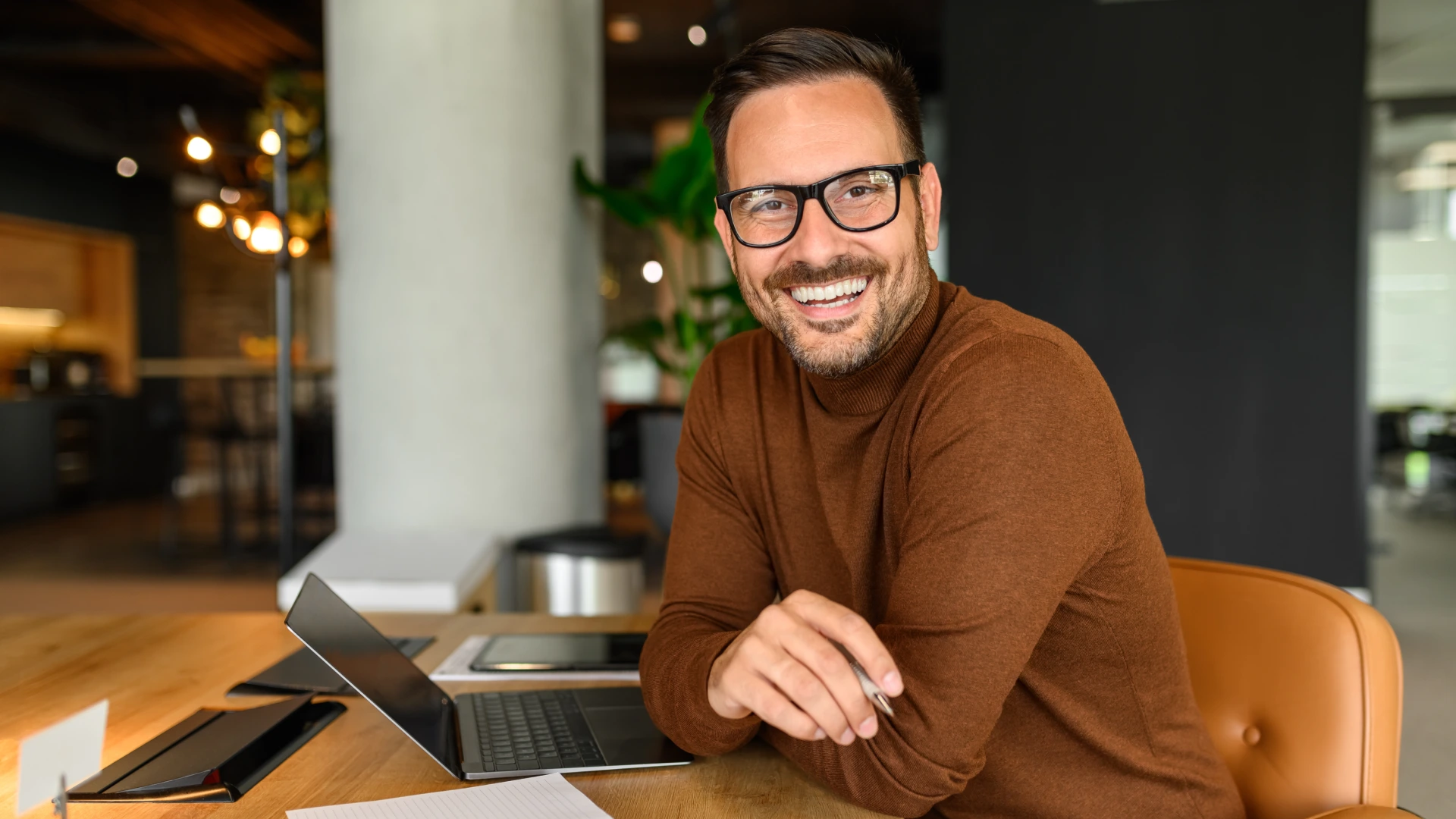Male freelancer with laptop and notepad on desk smiling at camera while sitting on chair